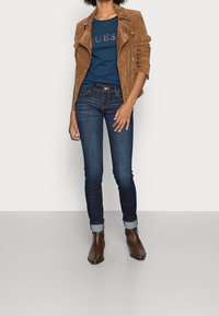 Kvinna som bär en brun mockajacka, en marinblå T-shirt med text, mörka uppkavlade jeans och bruna ankelskor som står mot en enkel bakgrund.