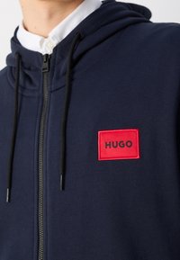 Sudadera con cremallera de color azul marino, con cordones negros y un parche rectangular rojo con la etiqueta "HUGO" en el lado izquierdo del pecho, llevada sobre una camisa blanca con cuello.