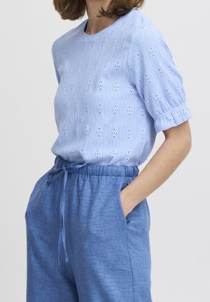 Hellblaues Oberteil mit Lochstickerei, kurzen Puffärmeln und rundem Ausschnitt, kombiniert mit hellblauen Leinenhose mit Kordelzug und seitlichen Taschen.
