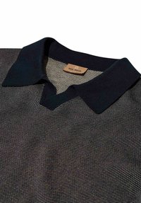 Detailný záber na čiernobiely pletený sveter s hladkým čiernym golierom a malou svetlohnedou štítkom s nápisom „MOS MOSH“ vo vnútri výstrihu.