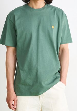 Vihreä puuvillapaita rentoa istuvuutta, jossa on pieni keltainen logo rintaosassa ja pyöreä kaula-aukko. Pehmeä, sileä tekstuuri.