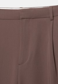 Gros plan sur la ceinture d’un pantalon marron sur mesure avec passants de ceinture et un détail de couture cachée sur la poche avant.