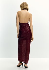 Nő áll h�áttal, burgundi színű, flitteres, pánt nélküli ruhát és fekete magas sarkú szandált visel egy fehér háttér előtt.