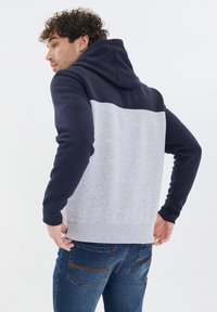 Grau Hoodie mit marineblauen Schultern, strukturierter Stoff, vordertasche im Känguru-Stil und gerippte Bündchen. Getragen mit blauen Jeans, die die Handposition zeigen.