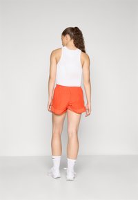 White ribbed sleeveless top paired with orange elastic waist shorts, featuring rolled hems. Model wears white socks and shoes.