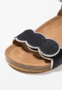 Černá semišová sandála s vlnitým páskem se stříbrným lemováním, texturovanou hnědou stélkou a odolnou gumovou podrážkou.