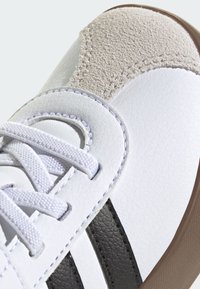 Zbliżenie na czubek białego trampka z beżową łatą z zamszu, czarnymi bocznymi paskami, białymi sznurowadłami i brązową, teksturowaną krawędzią podeszwy.