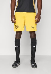 Gule fodboldshorts med sorte detaljer, der har et sort cirkulært logo og nummer 1. Båret med sorte sokker og sportssko.