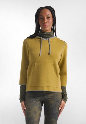 Vrouw met gevlochten haar die een mosterdgele hoodie draagt over een olijfgroene coltrui en donkere camouflagebroek, staand tegen een effen achtergrond.