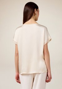 Crème zijde blouse met een losse fit, verlaagde schouders en een licht afgeronde zoom. Soepele textuur met korte mouwen en geen zichtbare sluitingen.