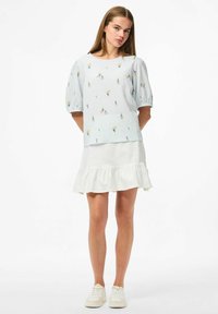 Top azzurro chiaro texturizzato con maniche a palloncino corte, caratterizzato da ricami floreali colorati, abbinato a una gonna bianca con volant e sneakers bianche.