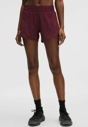 Lihaseline inimene, kes kannab tumepunaseid sportlustriksi, musti sokke ja musti jooksutrenne, seisab ühetaustal heleda tausta ees.