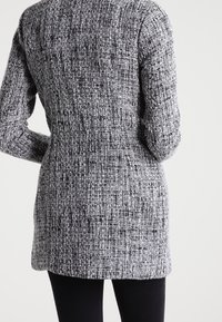 Cappotto lungo grigio e nero dalla texture, con una silhouette aderente, dotato di maniche lunghe e orlo dritto. Il materiale sembra tessuto e strutturato.