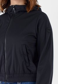 Giacca nera con cappuccio, dotata di zip frontale, vita elasticizzata e tasca laterale. Tessuto morbido con un logo in patch sulla manica.