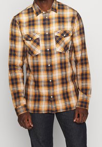 Uomo che indossa una camicia a scacchi gialla, blu navy e bianca con due tasche sul petto e jeans scuri.