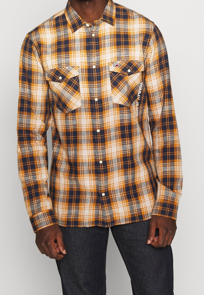 Uomo che indossa una camicia a scacchi gialla, blu navy e bianca con due tasche sul petto e jeans scuri.