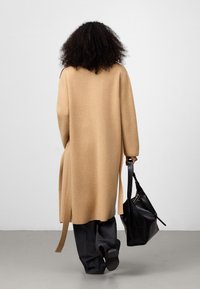 Cappotto lungo, beige, in lana con vestibilità rilassata e spacchi laterali, abbinato a pantaloni neri a gamba larga. Una borsa in pelle nera è tenuta in una mano.
