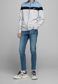 Felpa con zip di colore grigio chiaro, blu navy e blu con design a righe; abbinata a jeans blu e sneakers bianche chunky con accenti rossi.