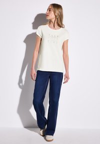 Cremefarvet t-shirt med korte ærmer og teksten "TAKE YOUR TIME", parret med mørkeblå vide jeans og lyse sneakers. Glat, afslappet design.
