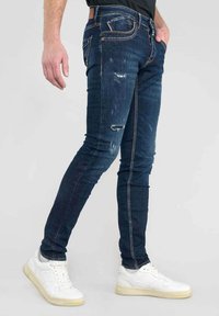 Jeans de denim azul de corte ajustado con detalles desgastados, cinco bolsillos y costuras en contraste, combinados con zapatillas deportivas blancas.