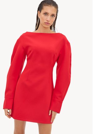 Femme vêtue d'une mini-robe rouge vif ajustée avec de longues manches volumineuses et un décolleté bateau, debout devant un fond uni.