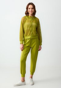 Limonengrüner Mesh-Reißverschlussjacke mit Kapuze, kombiniert mit passenden schmal geschnittenen Hosen mit Seitennähten und gerippten Bündchen. Weiße Sneaker.