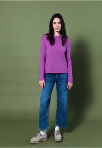 Frau, die vor einem olivgrünen Hintergrund steht und ein lila langärmliges Oberteil, blaue weite Jeans, weiße Socken und beigen klobigen Schuhen trägt.