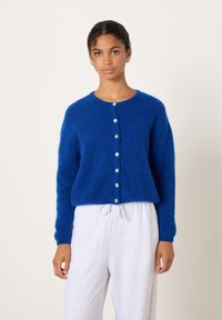 Jauna sieviete zilā pūkainā kardiganā ar pogām un gaiši pelēkās sporta biksēs stāv pret tīru balto fonu.