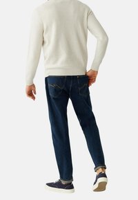 Pull en maille blanche, jean bleu foncé avec des coutures décoratives sur les poches arrière, et baskets bleu marine avec une semelle beige vues de dos.