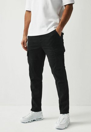 Black cargo pants with multiple pockets, slim fit, durable fabric, and contrasting stitching, paired with white sneakers.