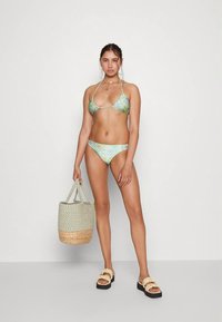 Biquíni tie-dye verde e azul com top de triângulo e parte de baixo de cintura baixa. Modelo carrega uma cesta de palha e veste sandálias de tiras claras.