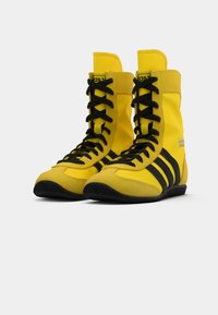 Yellow athletic boots with black laces and three black stripes. Made of durable material, featuring a high top design and textured sole.