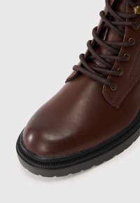 Bottine en cuir marron avec un bout rond, une couture saillante, des œillets en métal, et une semelle en caoutchouc noir avec des détails texturés.