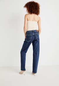 Ženska s kodrasto frizuro, oblečena v kremno majico brez rokavov, temno modre hlače z visokim pasom in bele sandale z višino, stoji ob beli ozadju.