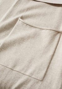 Tissu beige avec une texture douce présentant une poche carrée. Le design est simple avec des lignes épurées et un minimum de détails.