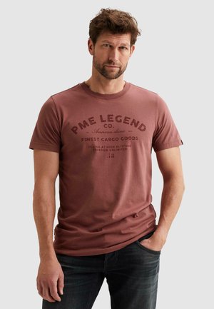 Uomo con capelli castani corti e barba, indossa una T-shirt grafica rossa sbiadita PME Legend e jeans scuri, mano in tasca.