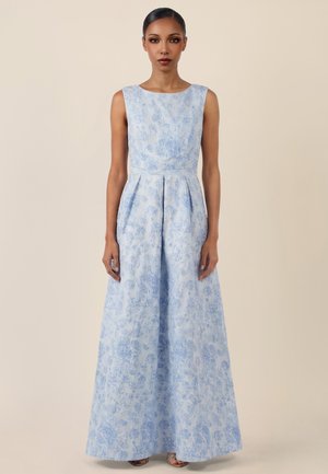 Frau trägt ein ärmelloses, hellblaues Abendkleid mit Blumenmuster und steht vor einem schlichten beigen Hintergrund.