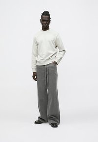 Pantaloni della tuta grigi oversize e felpa grigio chiaro realizzati in tessuto misto cotone. Modello in piedi con le mani nelle tasche, indossa scarpe nere.
