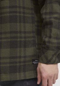 Grøn- og sortternet flannel skjorte med knapmanchetter, med en stikning i kanten og et mærke på siden til brandidentifikation.