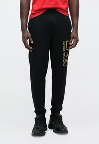 Fekete, slim fit szabású jogging nadrág, bal oldalon arany "Emporio Armani" logóval. Fekete sportcipővel és piros pólóval viselve.