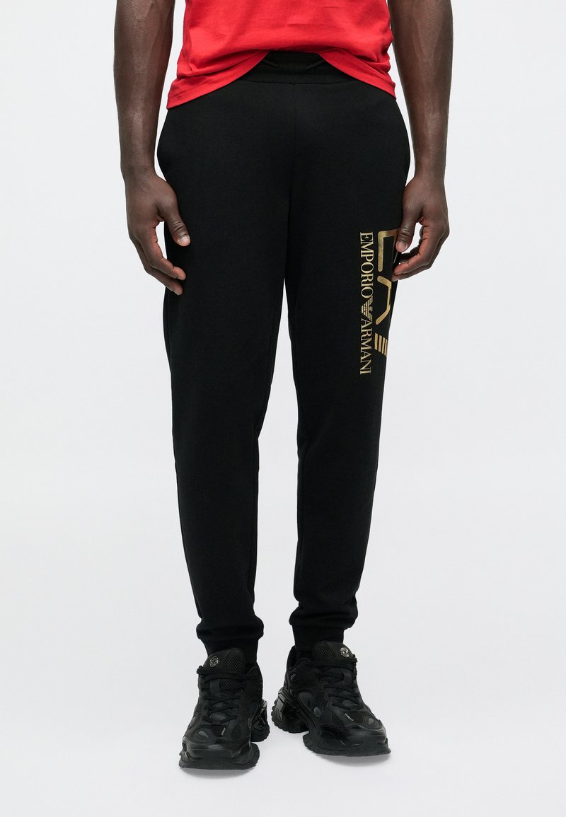 Fekete, slim fit szabású jogging nadrág, bal oldalon arany "Emporio Armani" logóval. Fekete sportcipővel és piros pólóval viselve.