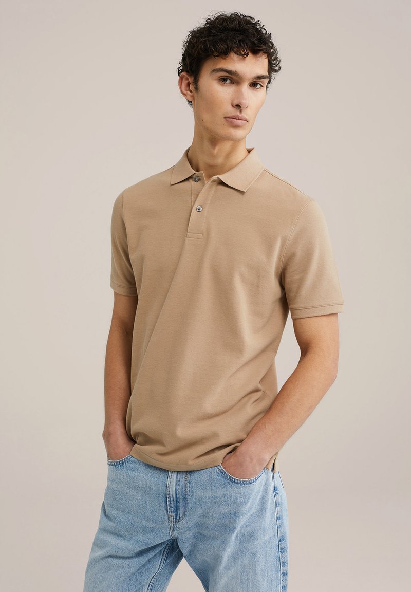 Camisa polo beige con cuello clásico, mangas cortas y una tapeta de tres botones, combinada con jeans de denim azul claro.