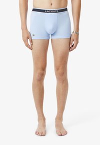 Boxer shorts de color azul claro, hechos de una tela suave, con una cinturilla negra que dice "LACOSTE" y un pequeño logo verde en el lado izquierdo.