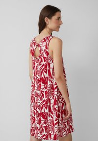 Kobieta ubrana w bezrękawnikową, czerwoną i białą sukienkę w abstrakcyjny wzór, z wycięciem na plecach związanym pod szyją, stojąca na jednolitym szarym tle.