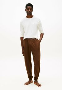 Giovane uomo in piedi a piedi nudi che indossa una camicia bianca a maniche lunghe e pantaloni marroni con coulisse, con le mani nelle tasche su uno sfondo bianco.