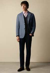 Completo maschile che include un blazer grigio-blu, gilet nero, camicia a righe rosa chiaro, jeans scuri e scarpe marroni. Design semplice con taglio su misura.