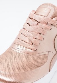 Närbild av en rosaguld Nike-sneaker med flätade snören, nättå och liten svart Nike-logotyp nära sulan på vit bakgrund.