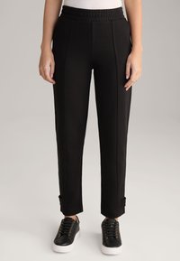 Zwarte trainingsbroek van een gestructureerde stof, met een elastische tailleband en taps toelopende benen, met zijnahts en sprakeen bij de enkels. Gecombineerd met zwarte sneakers.