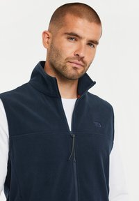 Navy fleece vest with a high collar, front zipper, and logo detail. Features a soft texture and sleek design, layered over a white shirt.