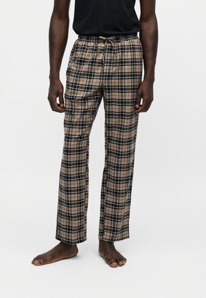 Mand iført beige og sort ternet pyjamasbukser med snører og en sort ærmeløs top, stående barfodet på en ensfarvet baggrund.